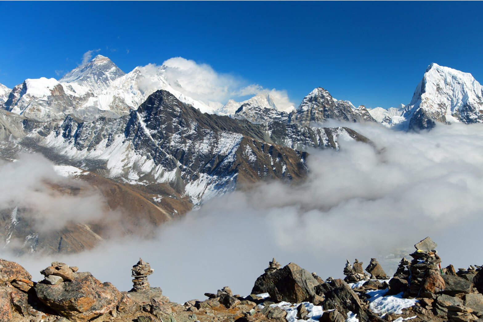 Mountain range with Mount Everest in the background with stacked stones in the foreground with blue sky at the upper levels and mist at the lower levels.