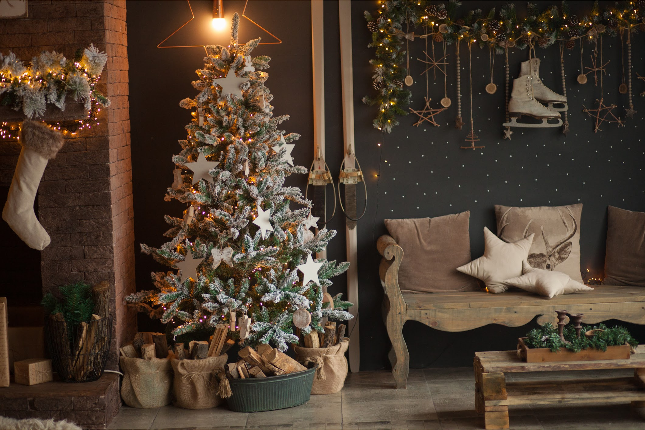 A room setting with a decorated Christmas tree with logs of wood below. The tree has wooden stars. There is a fireplace with a stocking. In the background there is a swag of foliage with some ice skates hanging down. There is a wooden bench with cushions .