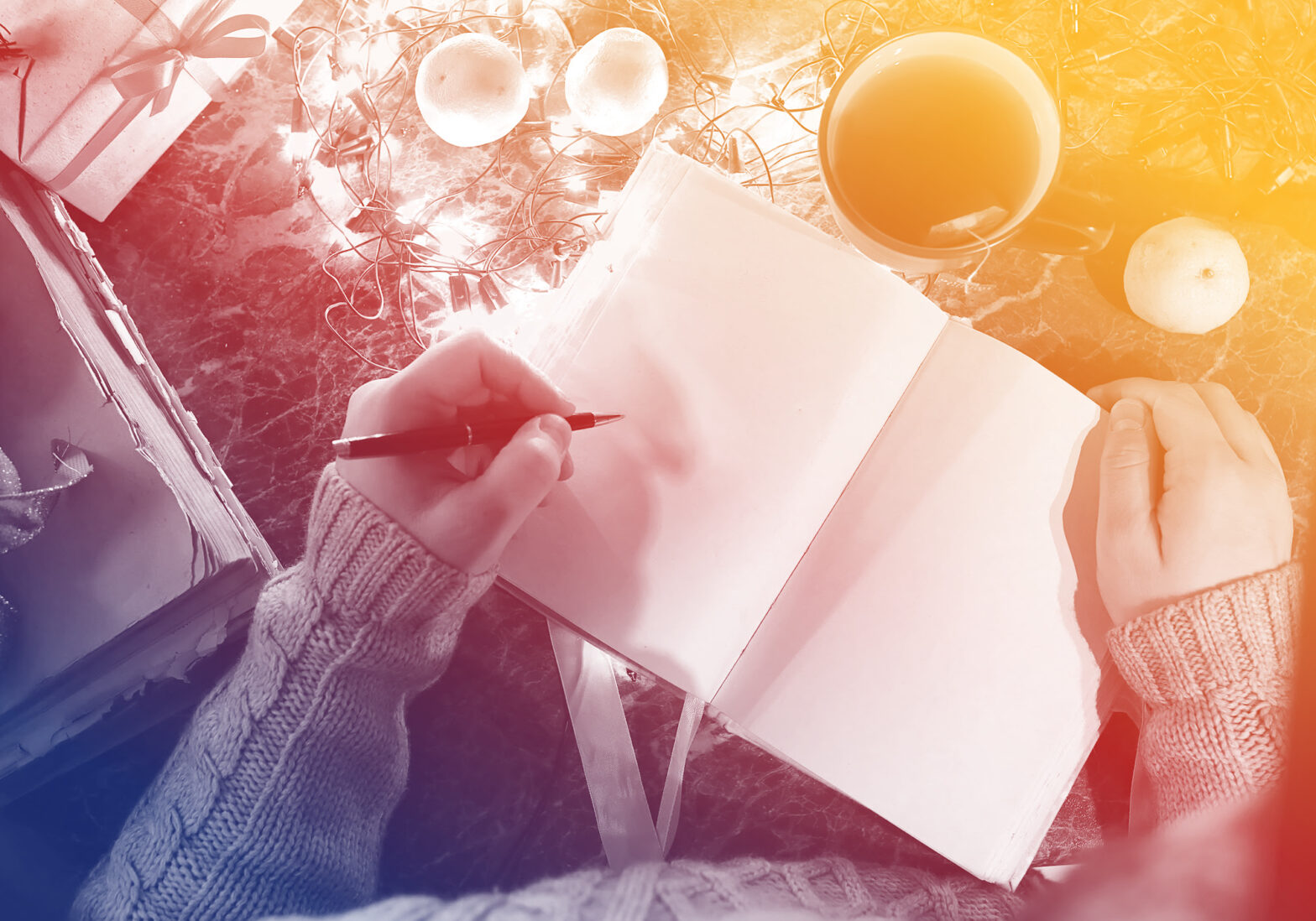 The arms and hands of a person holding a pen resting on an open blank notebook. In the surrounding area there appears to be a wrapped present, an old battered book, a mug of herbal tea and some fairy lights. There is an orange coloured glow from the top right hand corner of the page.