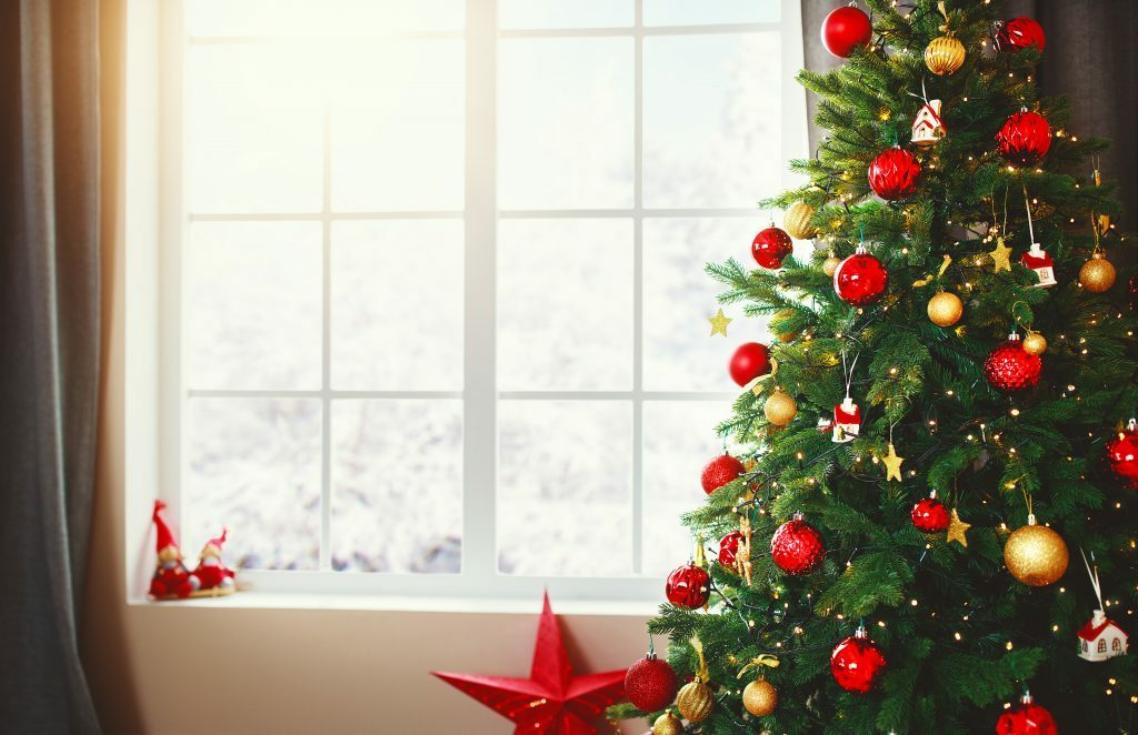 Window in the background and a decorated christmas tree in the foreground on the right hand side. Decorated with red and gold baubles . On the window sill are two gnome ornaments