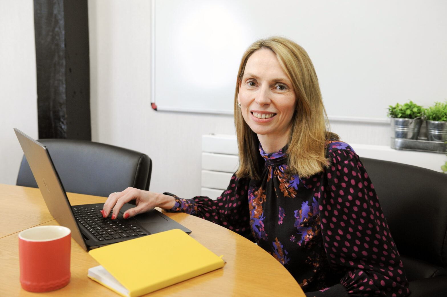 Lady with shoulder length hair in colourful top with laptop on desk and red cup and yellow book