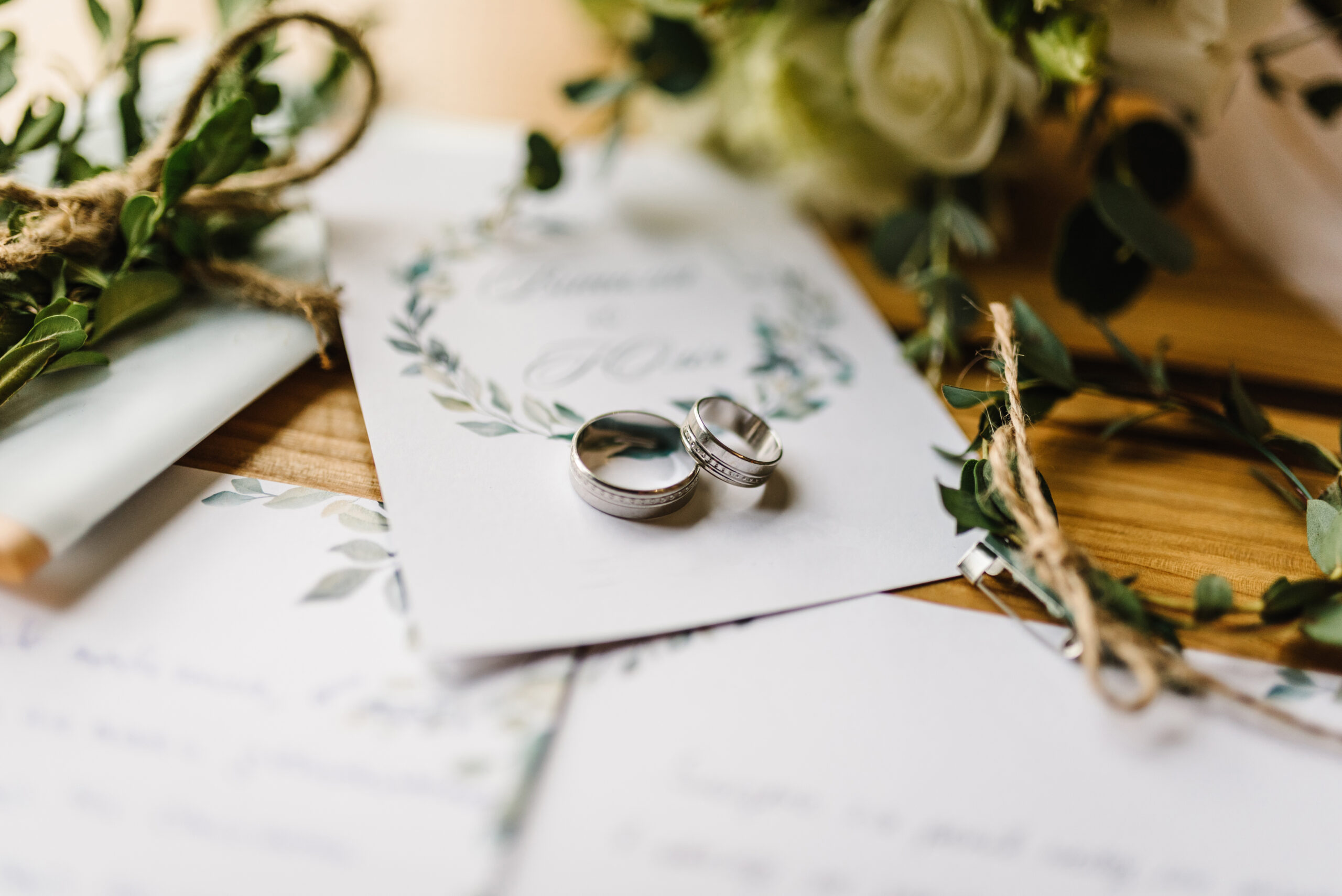 A larger and smaller ring resting on a blurred image of a wedding invitation. There are bundles of foliage on either side bound with jute string. Blurred in the background are some roses.