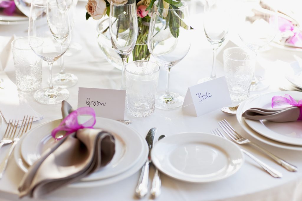 Table set for a wedding with place settings for the groom on the left and the bride on the right. There are plates, grey napkins with a dark pink ribbon, empty glasses, knives forks and a vase with flowers.