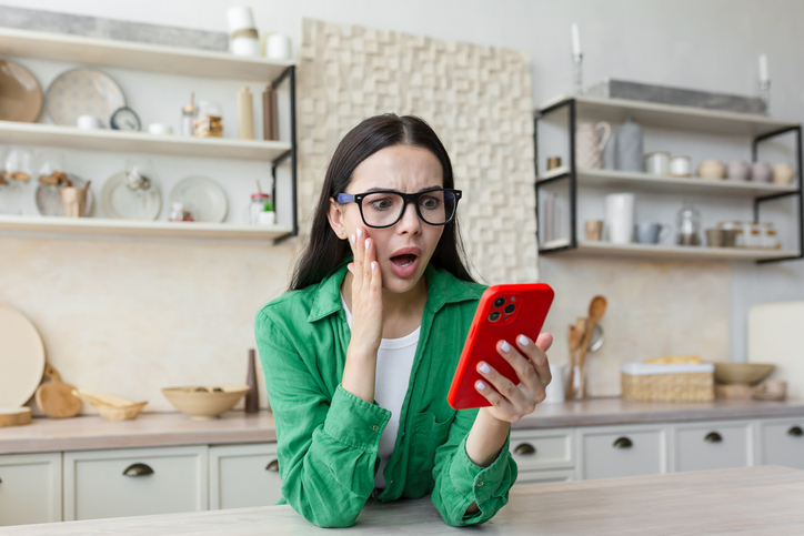 Lady with glasses and long brown hair wearing a green top sitting in a kitchen holding a red mobile phone. Her facial expression shows shock.