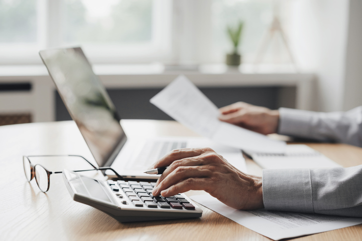 Blurred background image. In the foreground there is a desk with a laptop, calculator and pair of glasses. There is the hands and forearms of a man and he is holding a pen as he touches the calculator and he is holding some papers.