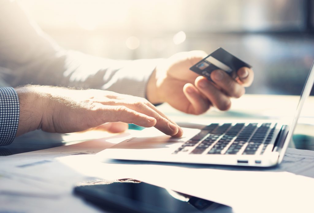 Hands of a person holding a credit card with sunlight shining through. They are using the touch pad of a laptop.