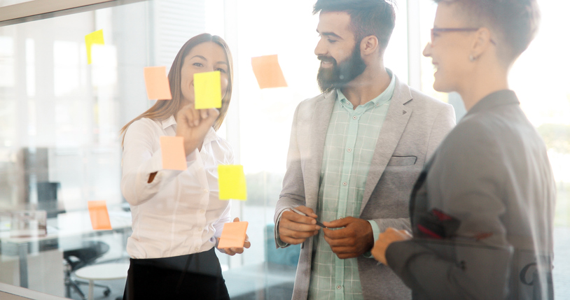 Business people gathered by a glass board with square sticky notes stuck on it.